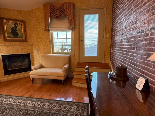 A cozy living room with brick wall, fireplace, window bay, patterned chair, and a small round table in warm, classic decor.