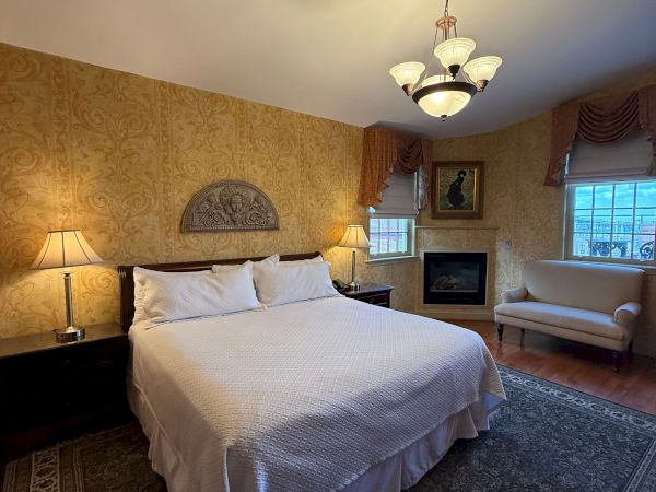 A warmly lit bedroom with a four-lamp chandelier, patterned wallpaper, bed, sofa, fireplace, and large window.