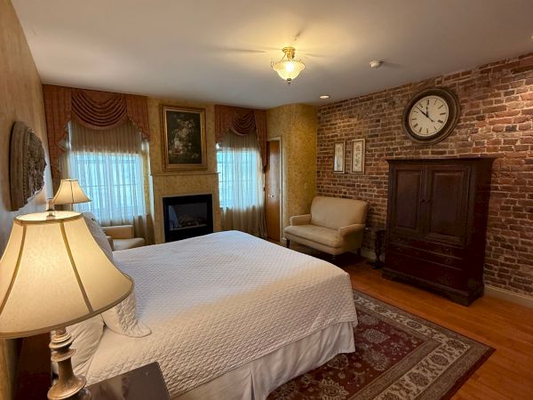 A cozy bedroom with a bed, bedside lamps, brick fireplace wall, mantel decor, framed pictures, and a wall clock.