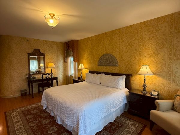 A cozy hotel bedroom with a chandelier, king bed, bedside lamps, patterned wallpaper, curtains, and a vanity mirror area.