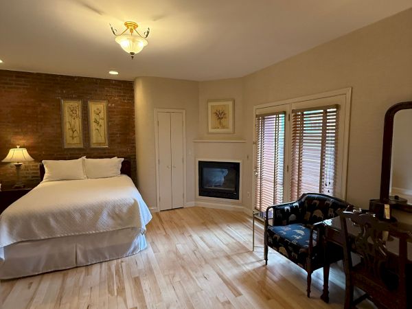 Cozy bedroom with brick accent wall, lit chandelier, fireplace, bed, floor-to-ceiling blinds, and a patterned armchair.