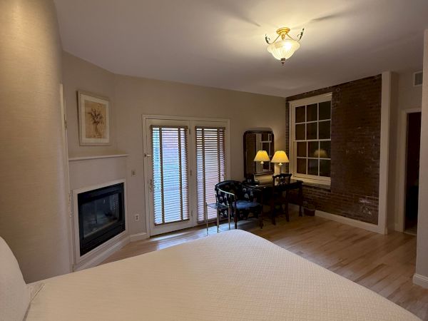 A cozy living room with a ceiling chandelier, brick accent wall, fireplace, blinds-covered windows, and two table lamps.