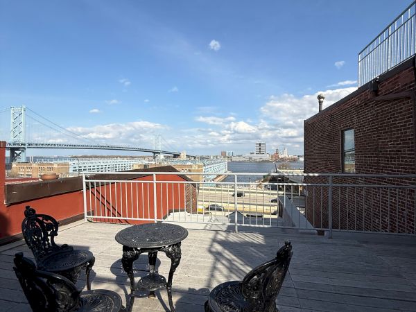 A rooftop terrace with ornate chairs, overlooking a wide river, a large steel bridge, and a city skyline under blue skies.