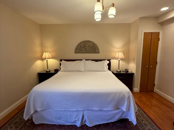 A neatly made hotel-style bedroom with a bed, ceiling light, lamps, dresser, and wooden door.