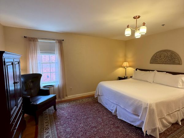 A cozy bedroom with a bed, chandelier, wall lamp, patterned rug, curtains, and a chair by the window.