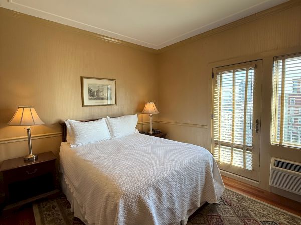 A cozy bedroom with a neatly made bed, two bedside lamps, patterned rug, a ceiling chandelier, and large window blinds.