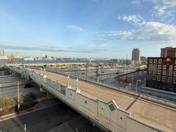 A riverfront city view with cranes, construction rubble, busy roads, and a bright blue cloudy sky.