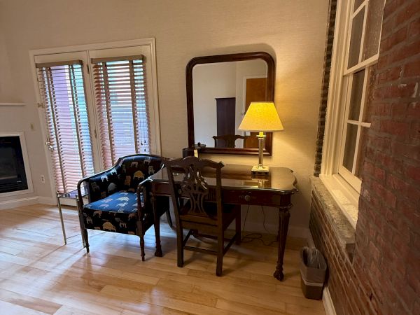 A cozy corner office with a desk and chair, a lit table lamp, a wall mirror, blinds-covered windows, and exposed brick walls.